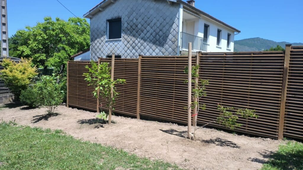 Clôture en bois ajouré et jeunes arbres dans un jardin avec vue sur les montagnes et une maison.
