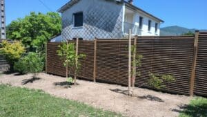 Clôture en bois ajouré et jeunes arbres dans un jardin avec vue sur les montagnes et une maison.